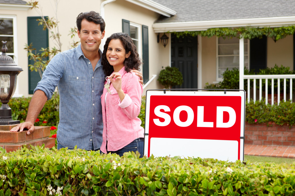 couple smiling with woman holding house key in front of a sold house